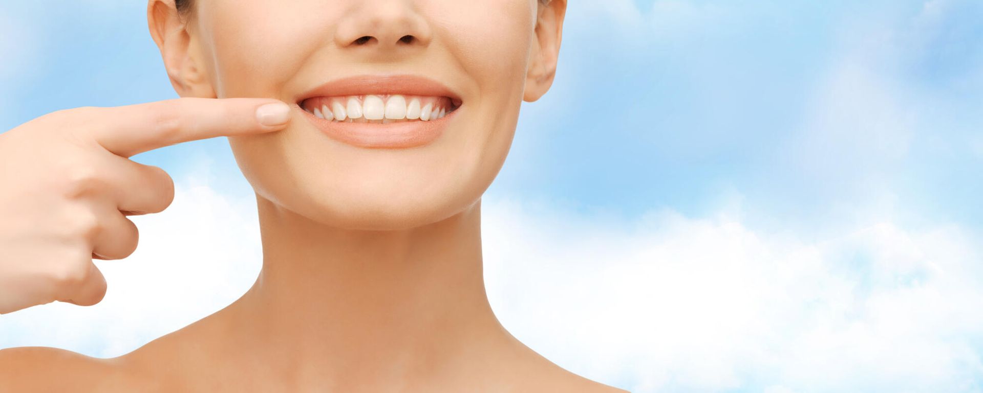 Woman smiling, pointing to her teeth, against a blue, cloudy background.