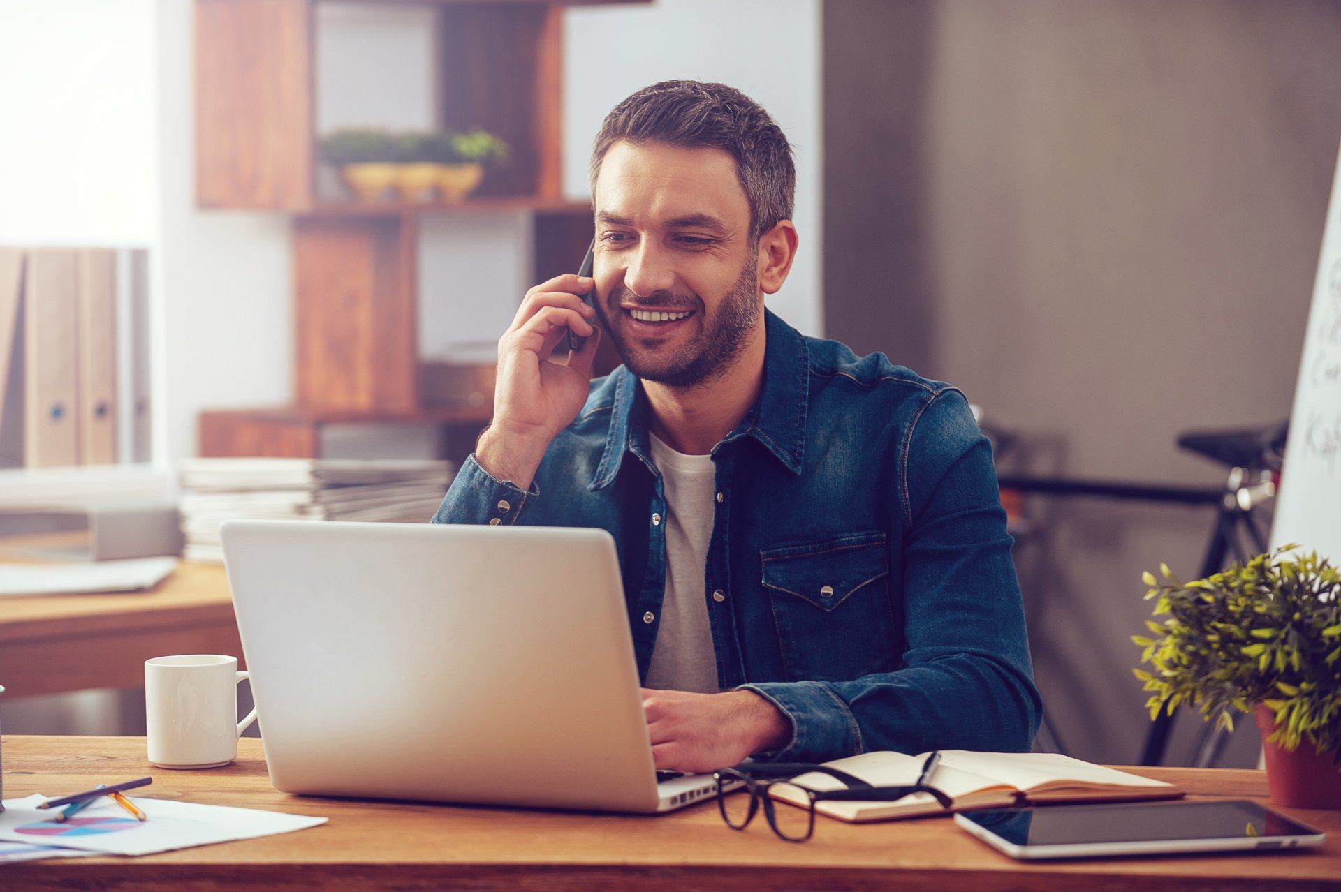 A man is sitting at a desk with a laptop and talking on a cell phone.