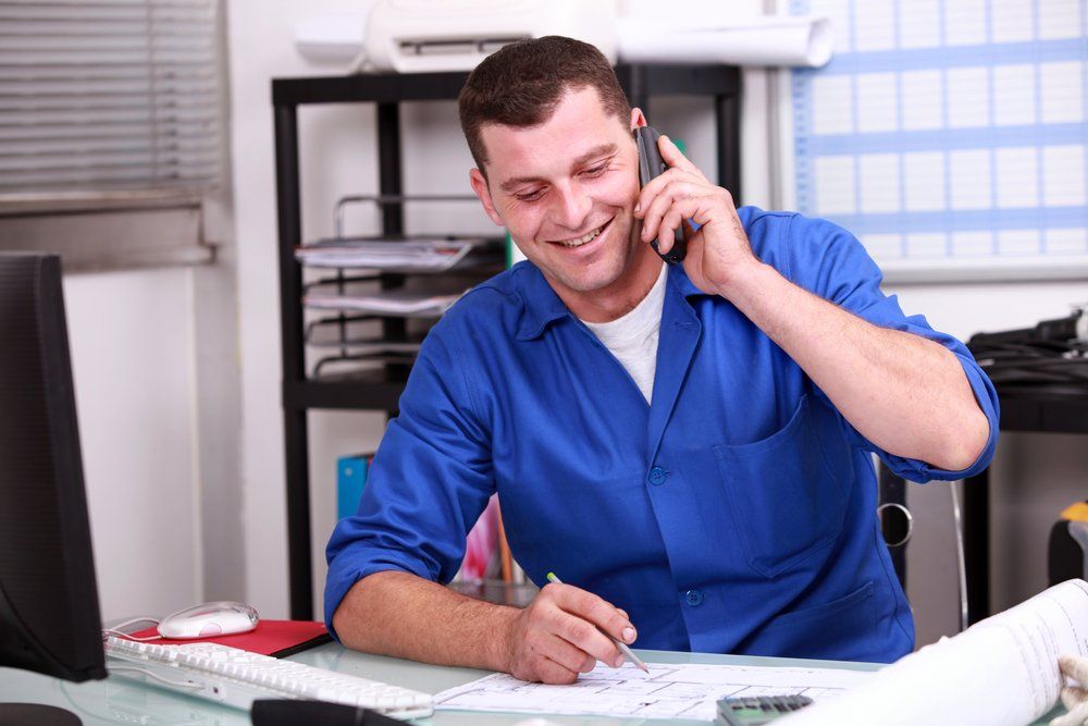 A man in a blue shirt is sitting at a desk talking on a cell phone.