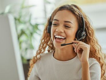 A woman is wearing a headset and smiling while talking on a computer.