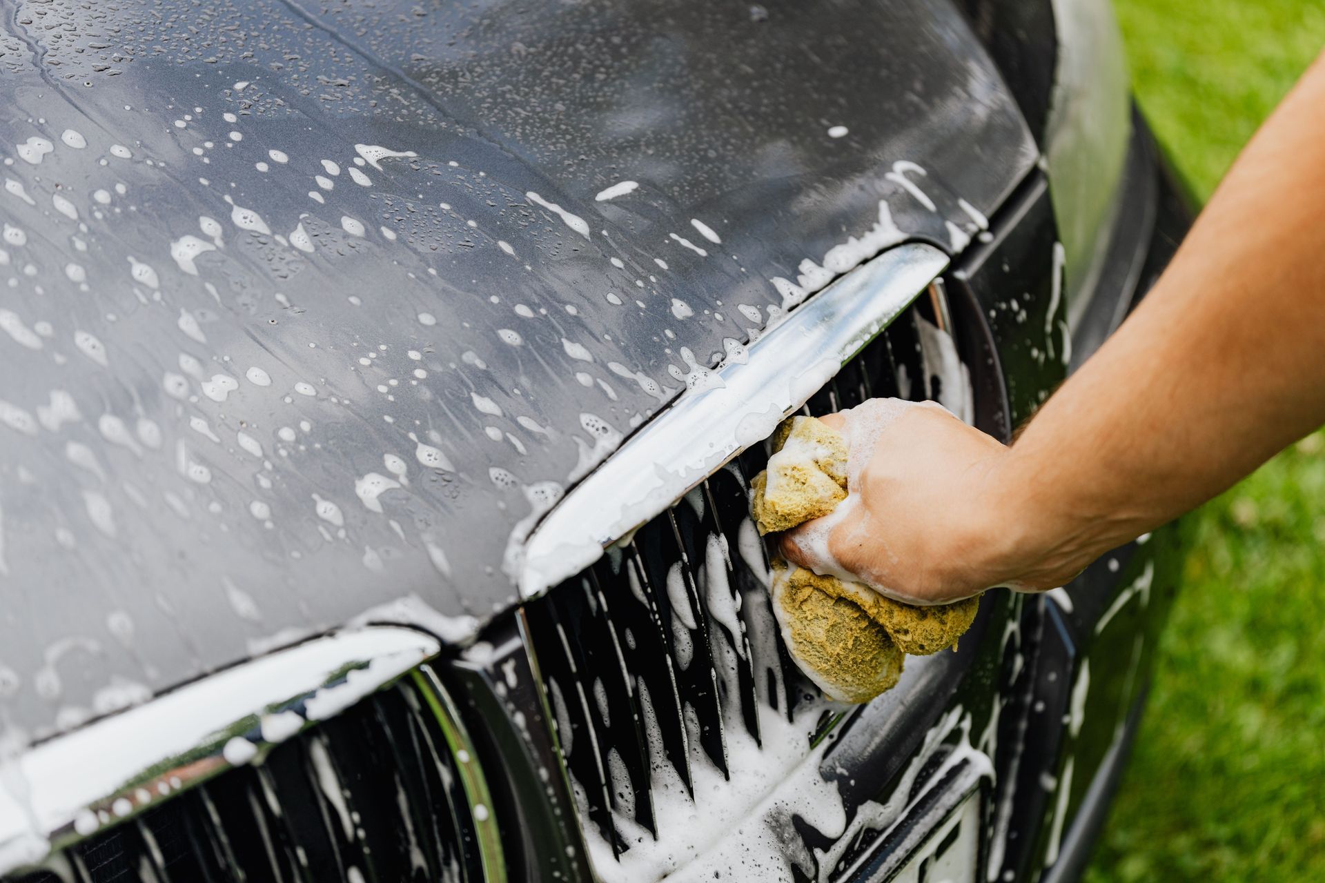 Hand washing a black car’s front grille with a yellow sponge and soapy suds
