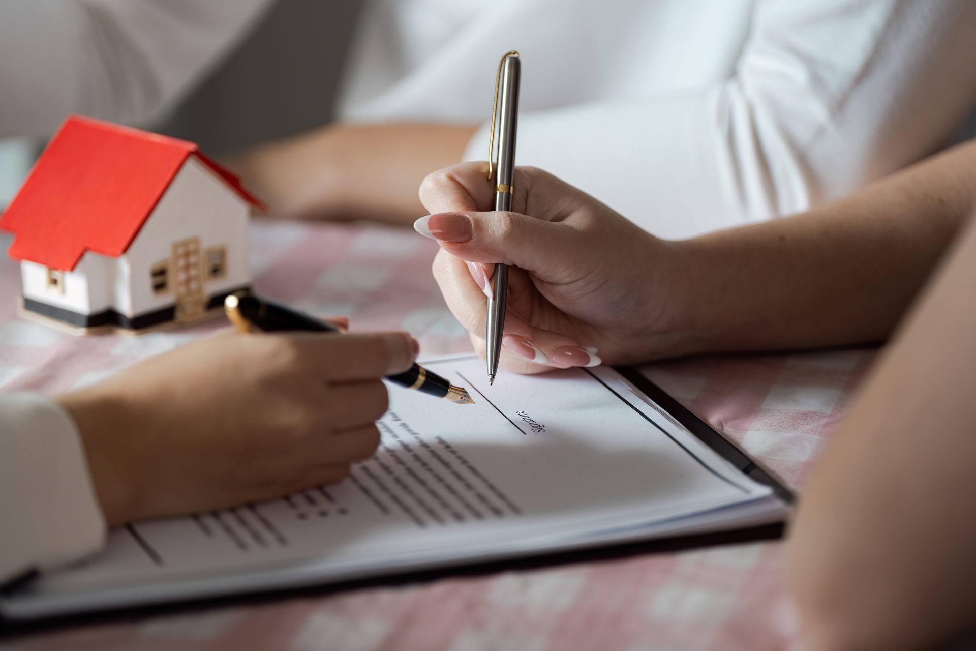 Person signs a real estate document beside a small model house on a desk.