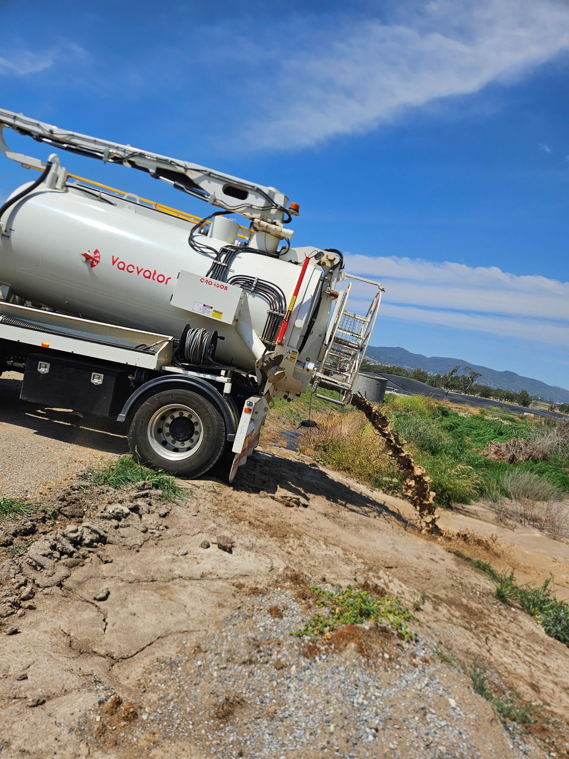 White truck dispensing brown liquid onto dirt ground with a field and mountains in background.