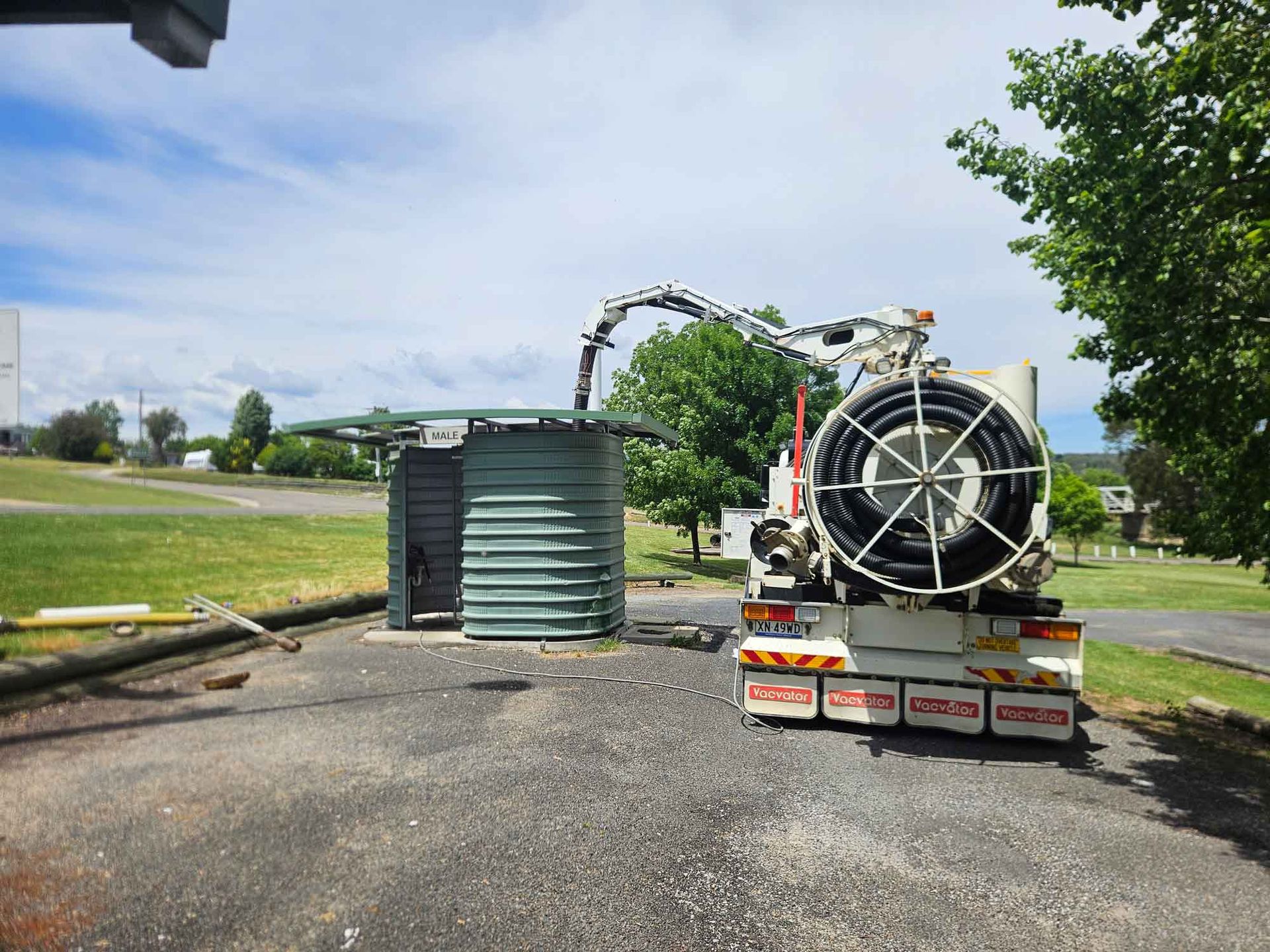 White truck with raised tank, dumping contents onto ground, worker nearby. Blue sky, green grass.
