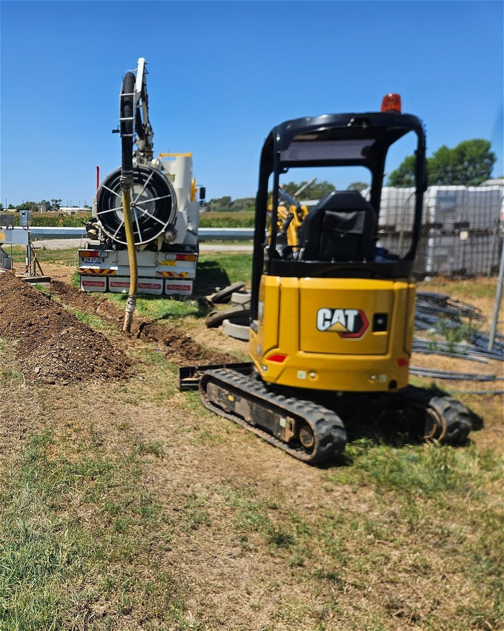 A yellow Caterpillar mini excavator parked on a grassy field next to a utility truck with a large hose reel.