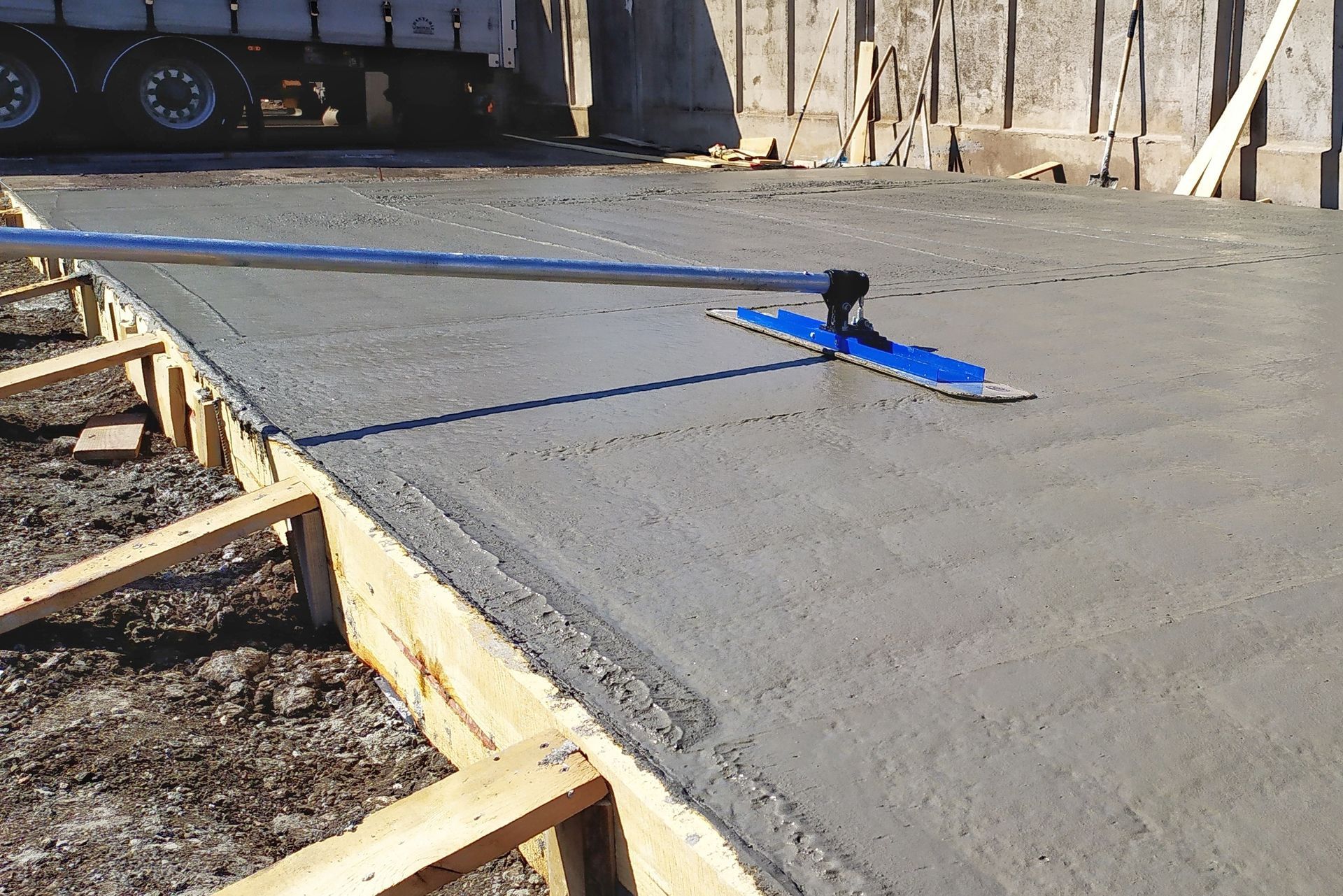 Béton fraîchement coulé nivelé à l'aide d'un long outil bleu dans un cadre en bois ; chantier de construction.