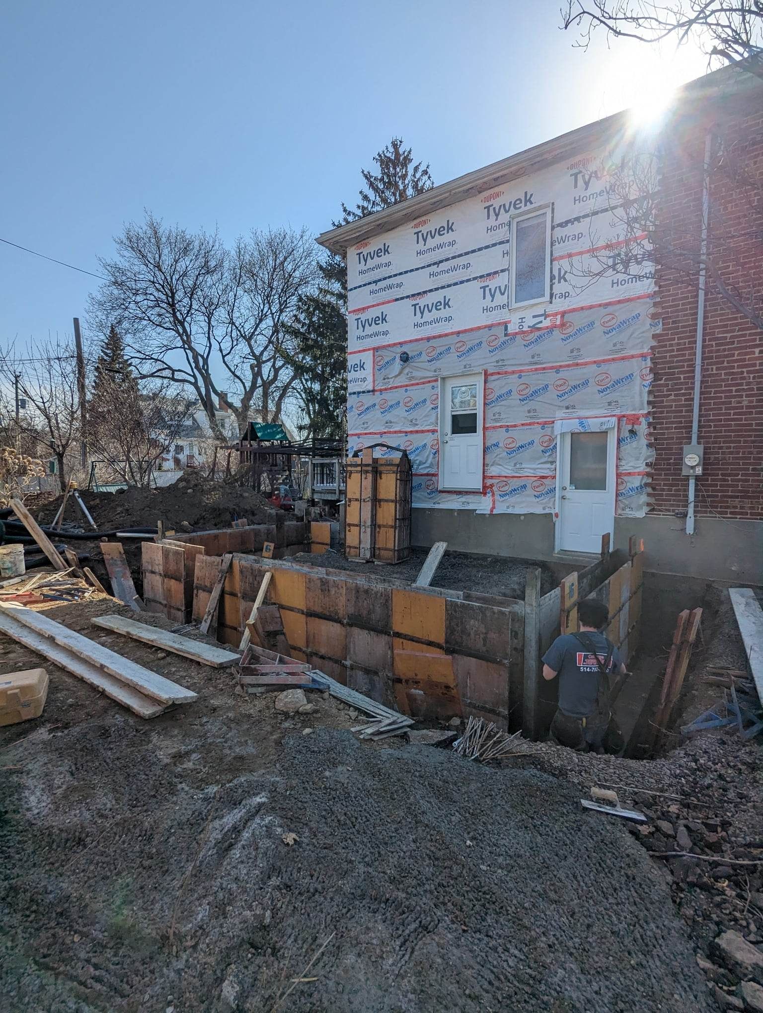 Chantier de construction avec coffrages en bois pour les fondations, ouvrier dans un trou, soleil radieux.