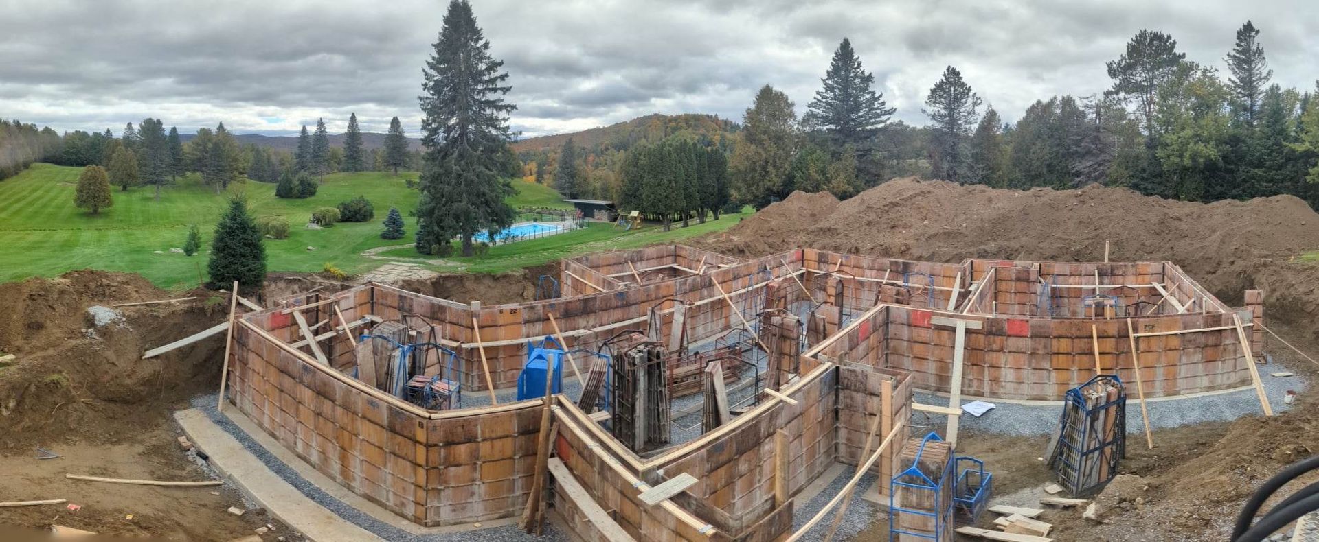 Chantier de construction : coffrages en bois pour les fondations d’un bâtiment, avec des ouvriers, sur une colline verdoyante sous un ciel couvert.