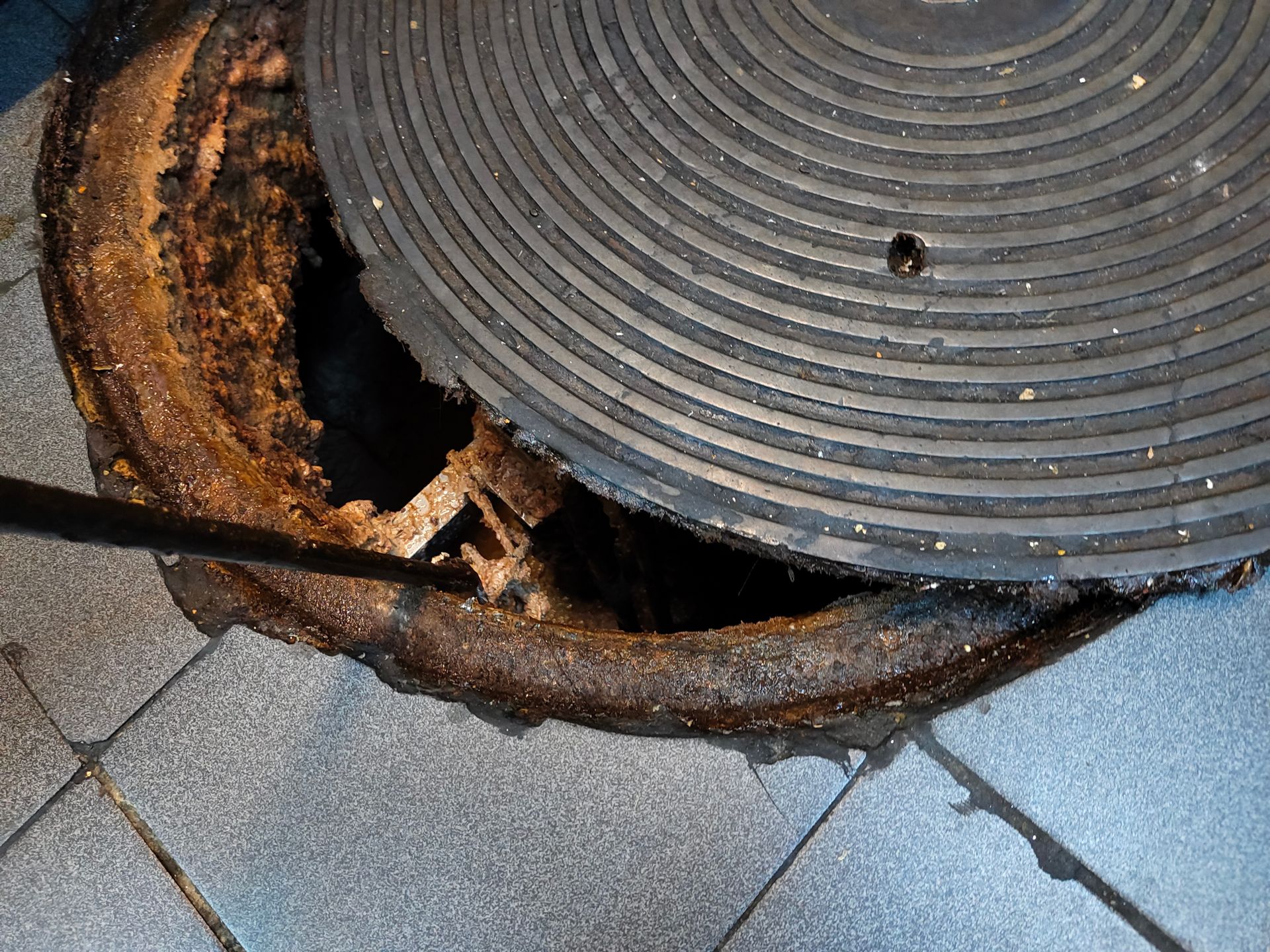 Severely rusted manhole cover with broken asphalt on a paved surface