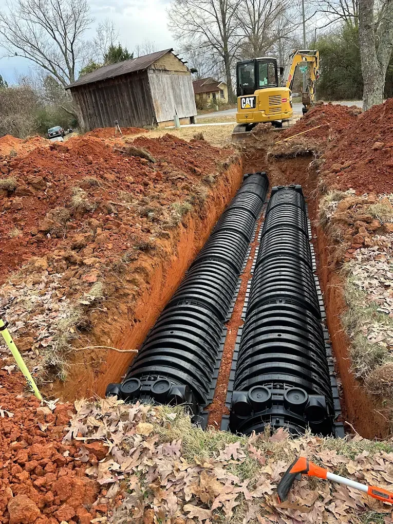 Excavator digging a trench with black drainage pipes beside a small shed in a muddy yard