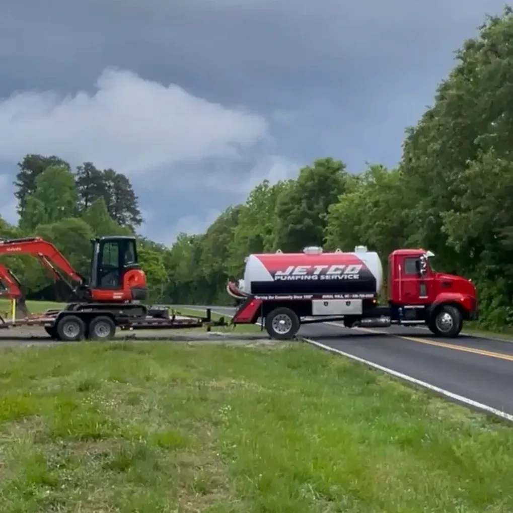 Red Jetvac vacuum truck and orange excavator on a roadside near trees under cloudy skies