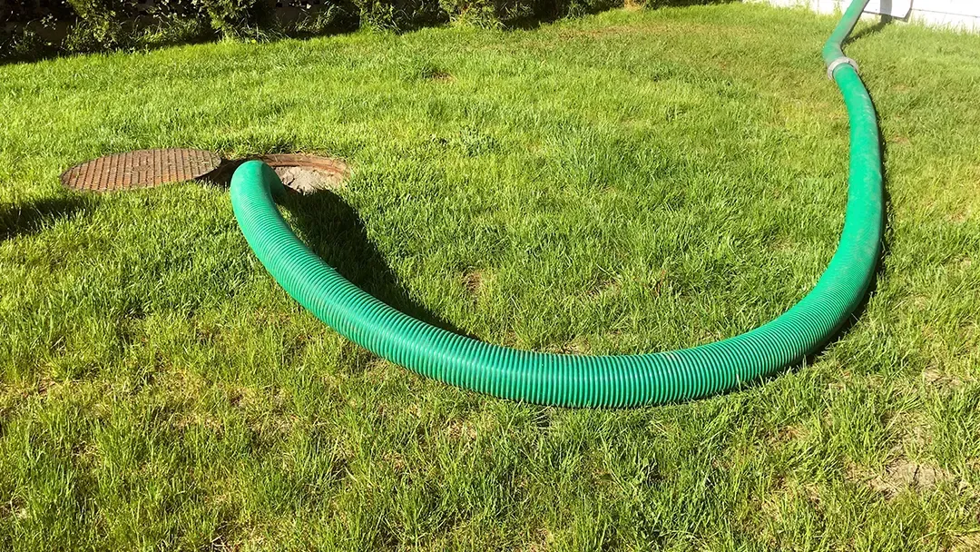 Green corrugated hose coiled across a grassy lawn beside a drain grate.