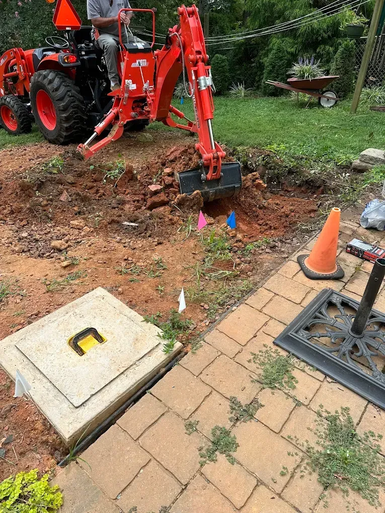 Orange backhoe digging near a patio, with a utility cover and traffic cone in the foreground