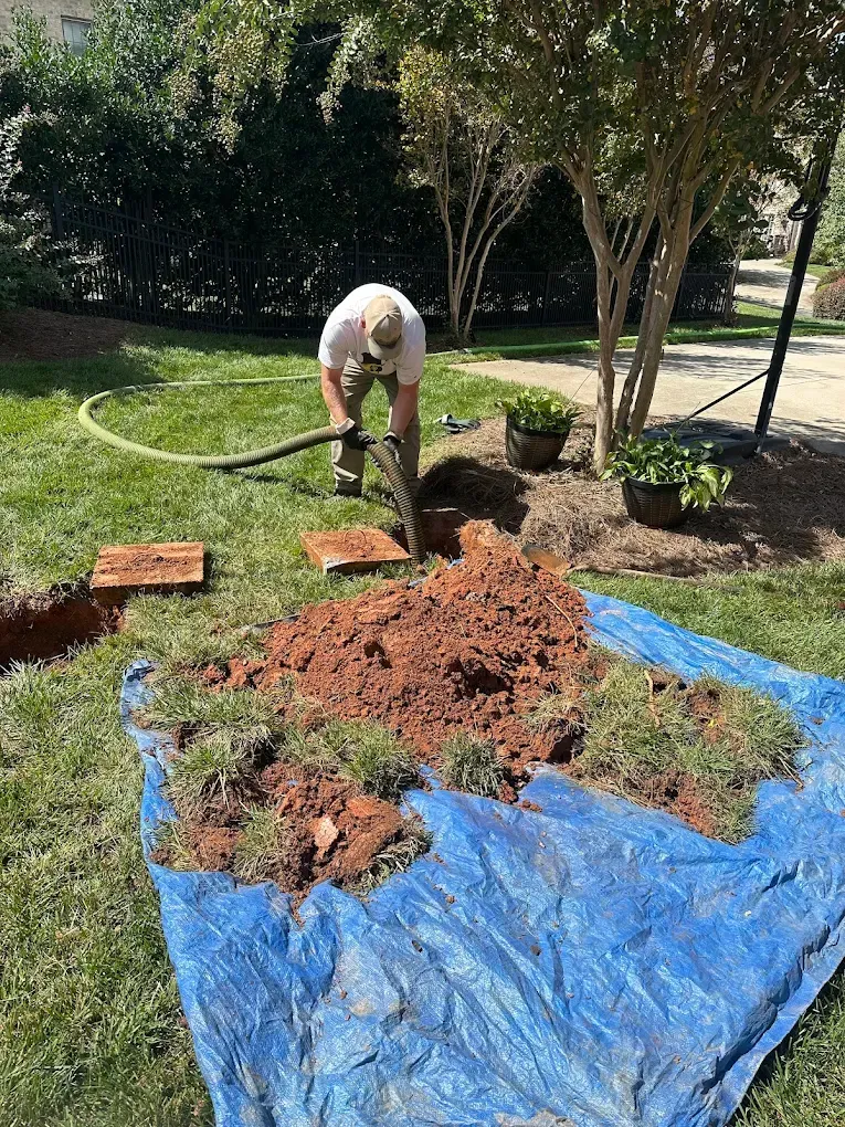 Person raking soil beside a blue tarp in a garden with trees and potted plants