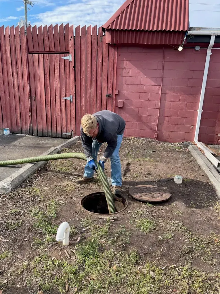 Person using a hose to pump or drain a round hole beside a red wooden shed and fence.