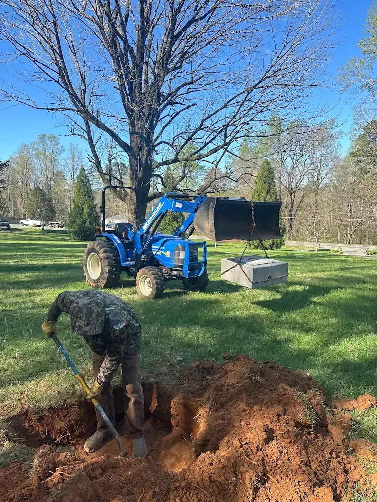 Person shoveling dirt in yard beside blue tractor and large hole