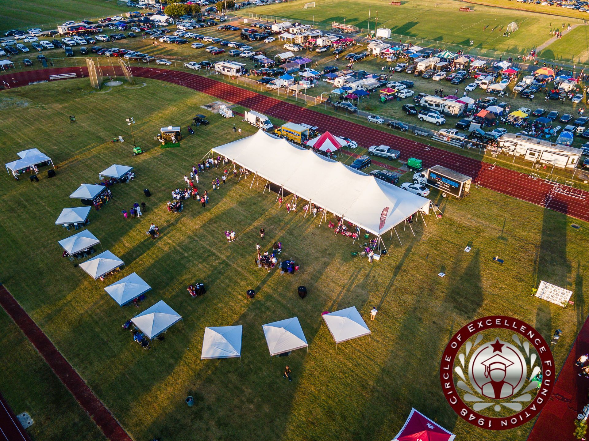 Aerial view of outdoor event with tents, vehicles, and a large white tent on a grassy field.