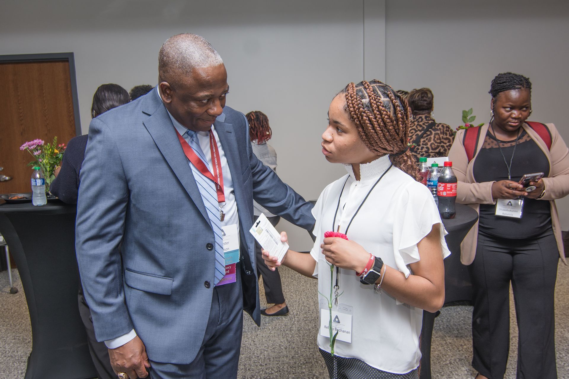Man in suit and woman in white top conversing at an event; other people and refreshments in background.