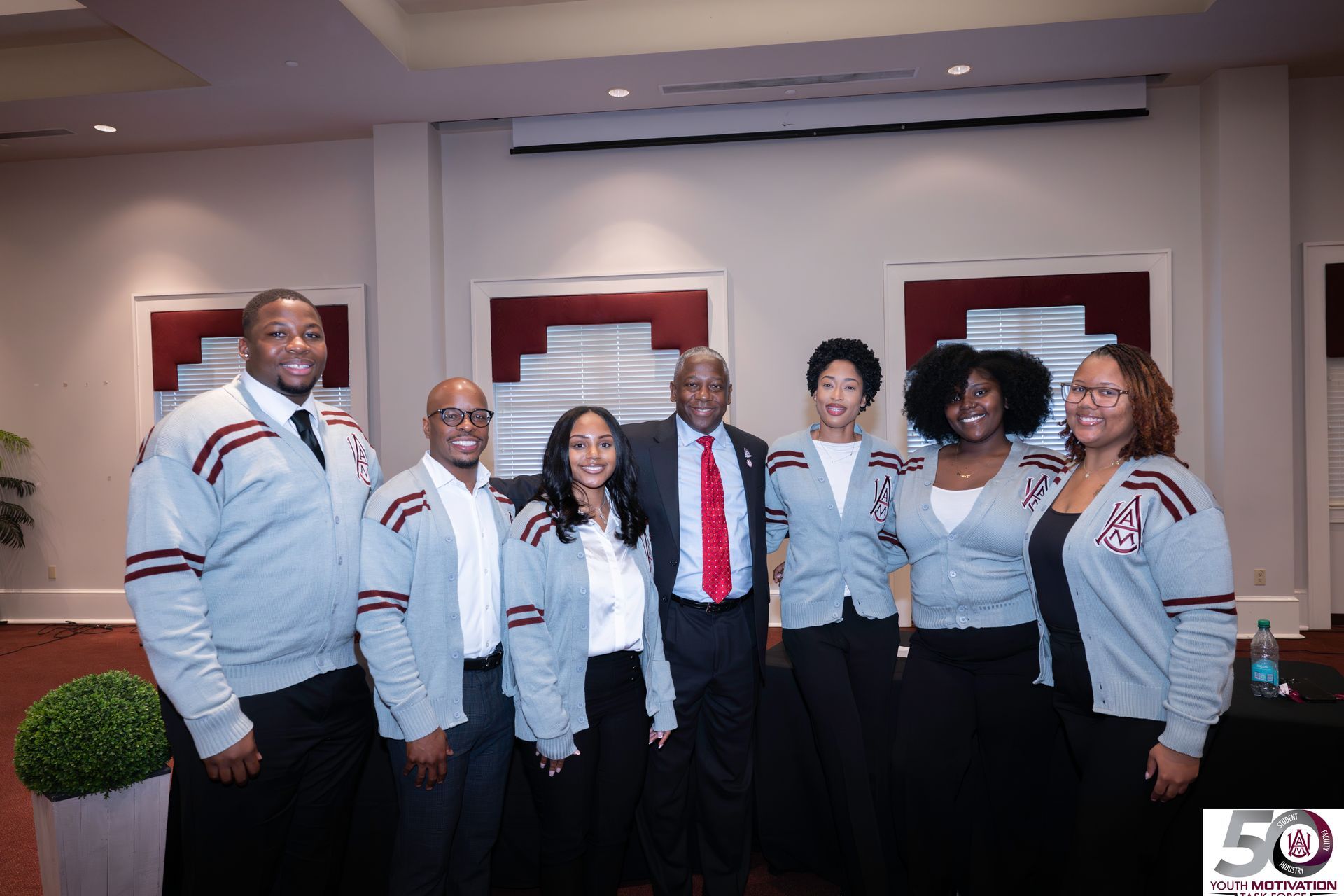 Group of people in matching sweaters pose with a man in a suit, in a room with a presentation screen.