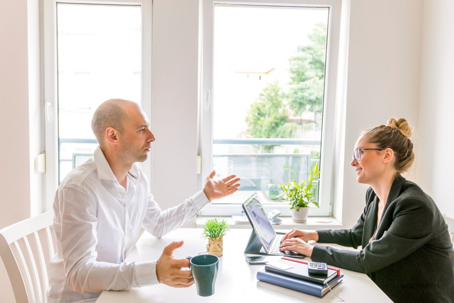 Two people talking at a bright office table with a laptop and notebooks