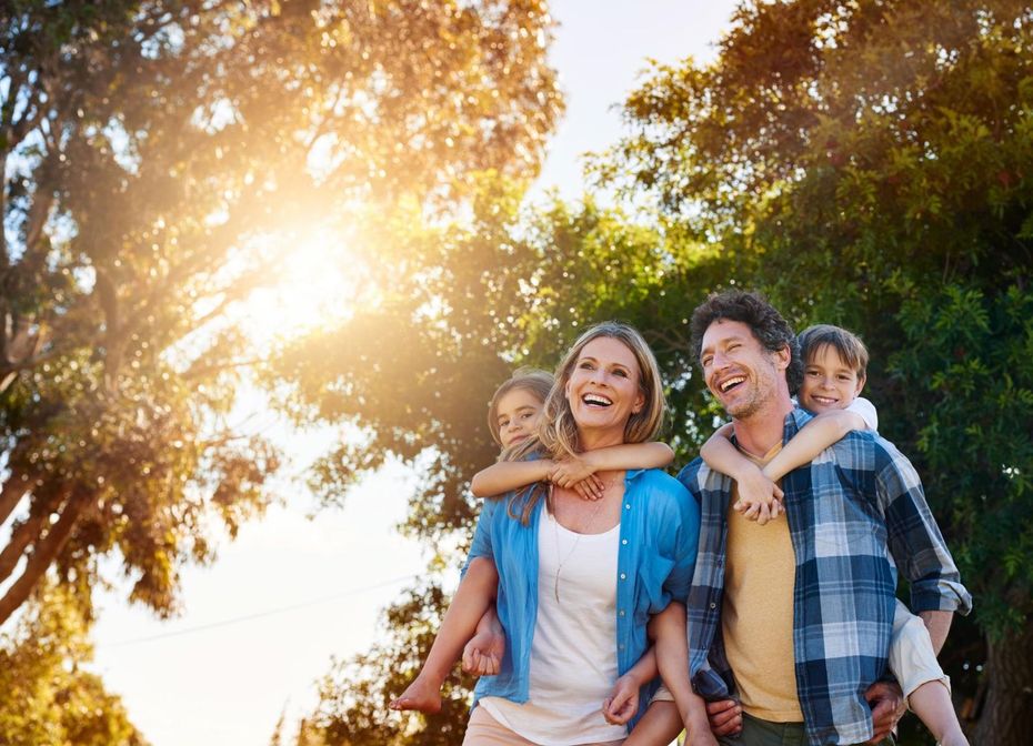 Smiling family outdoors in a sunny park, with a child on a man's shoulders and warm sunlight through trees