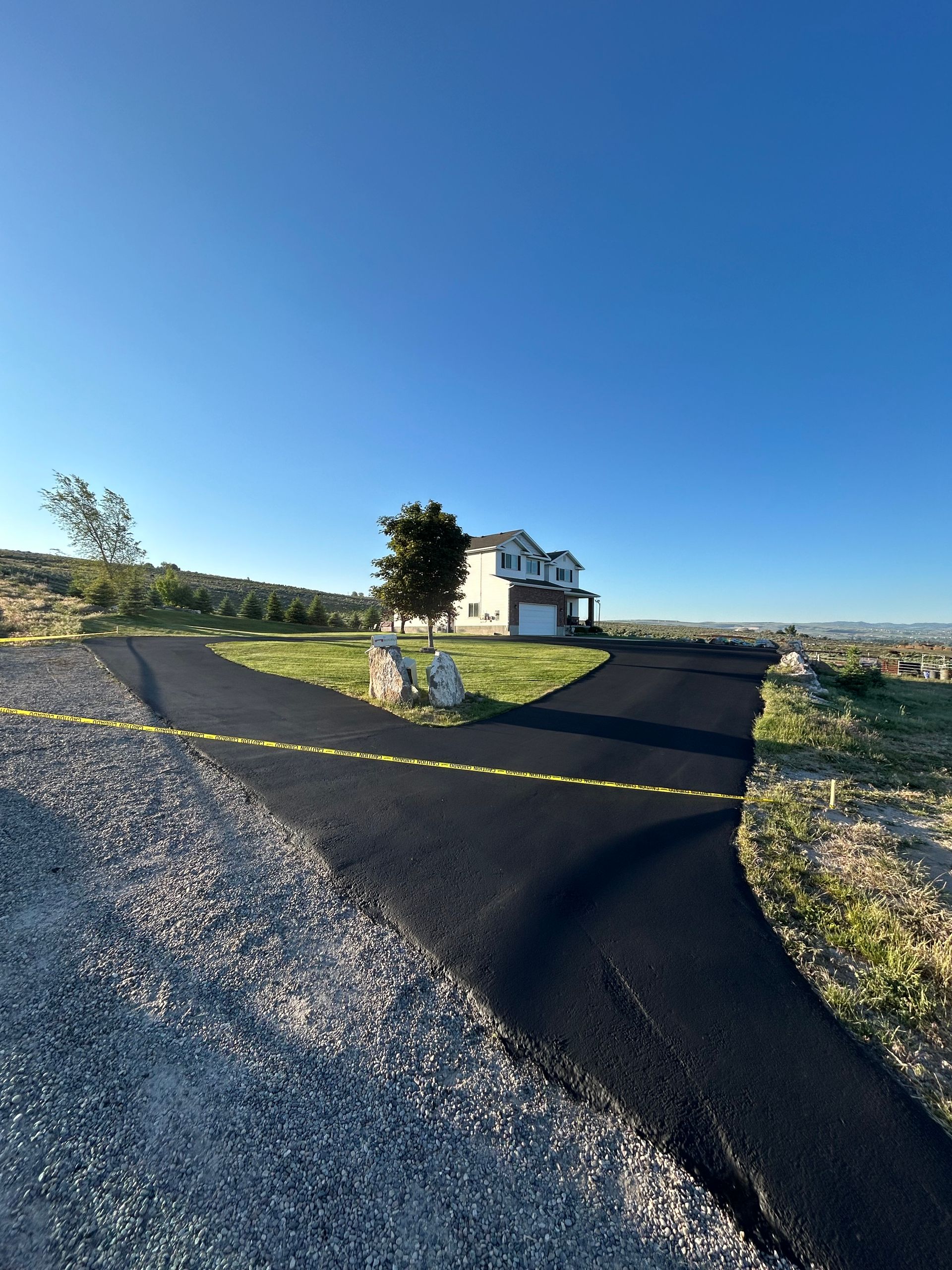 A split asphalt driveway leads up a grassy slope toward a two-story residential house under a clear blue sky.