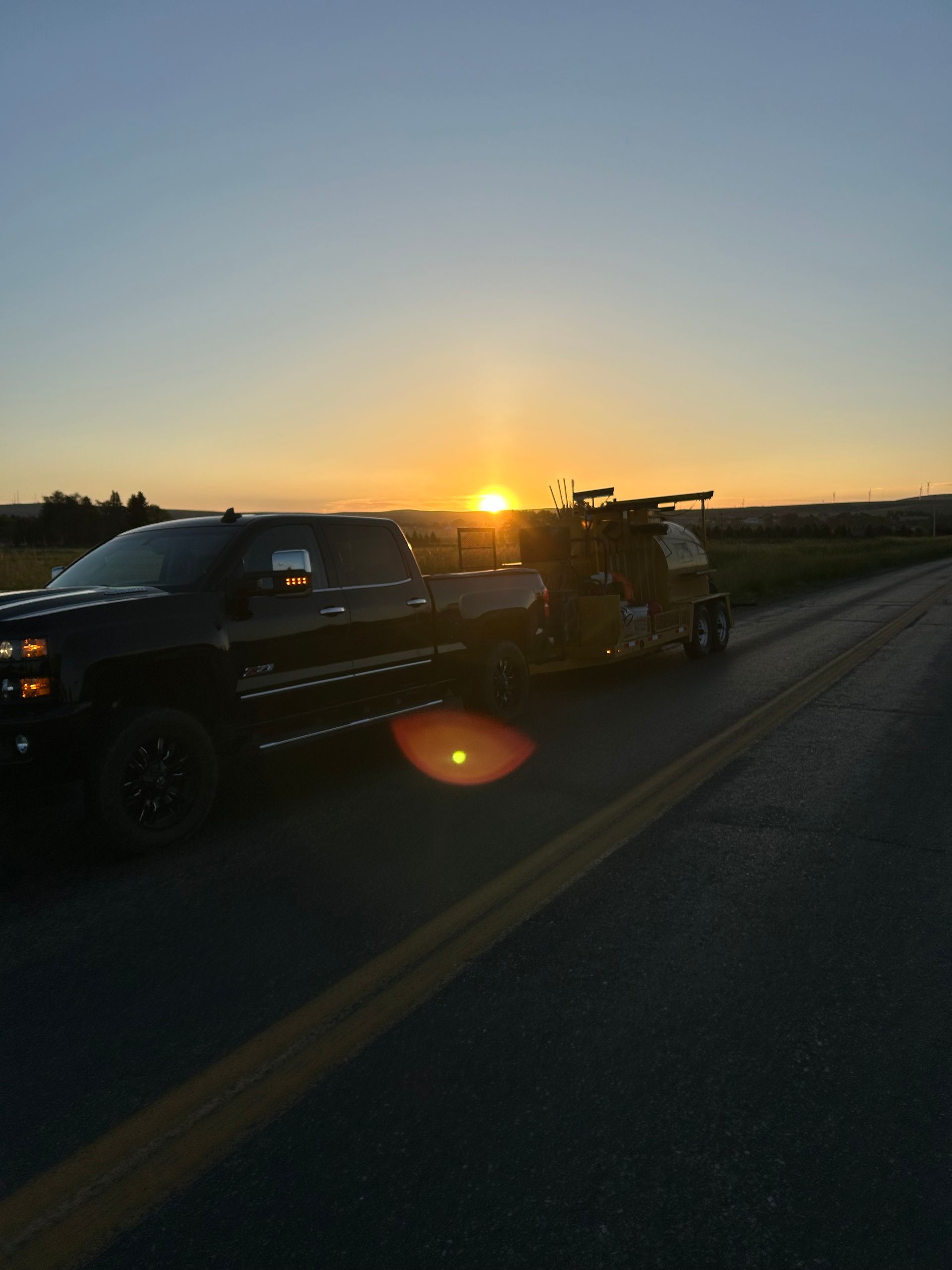 A black pickup truck towing a trailer on a paved road during a golden sunset.