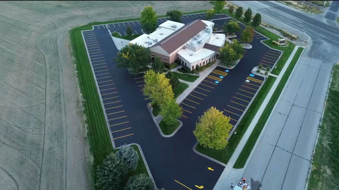 Aerial view of a commercial building with a freshly paved asphalt parking lot surrounded by green trees and fields.