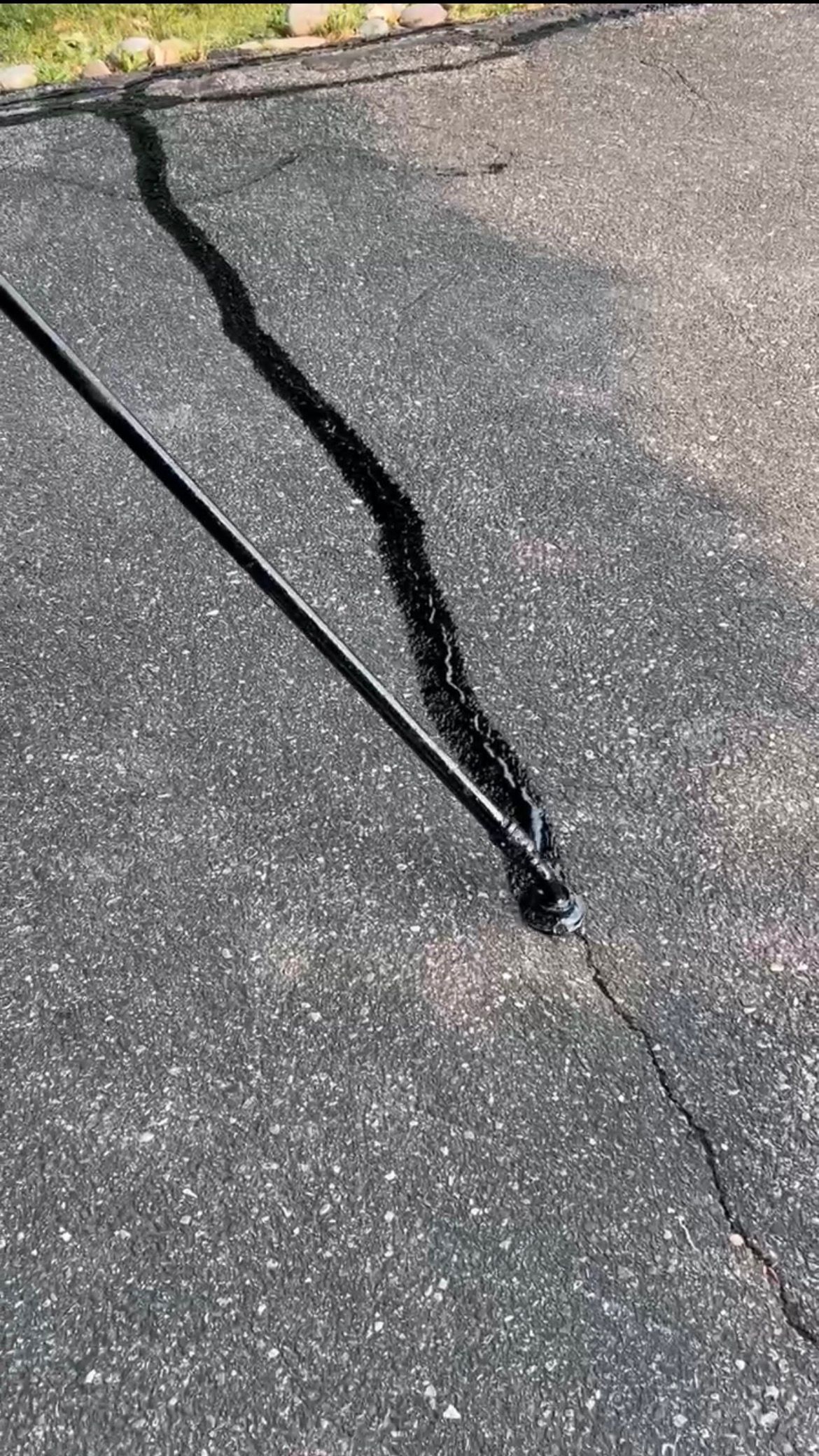 A dark tool with a long handle applies black liquid sealant to a crack in an asphalt driveway.