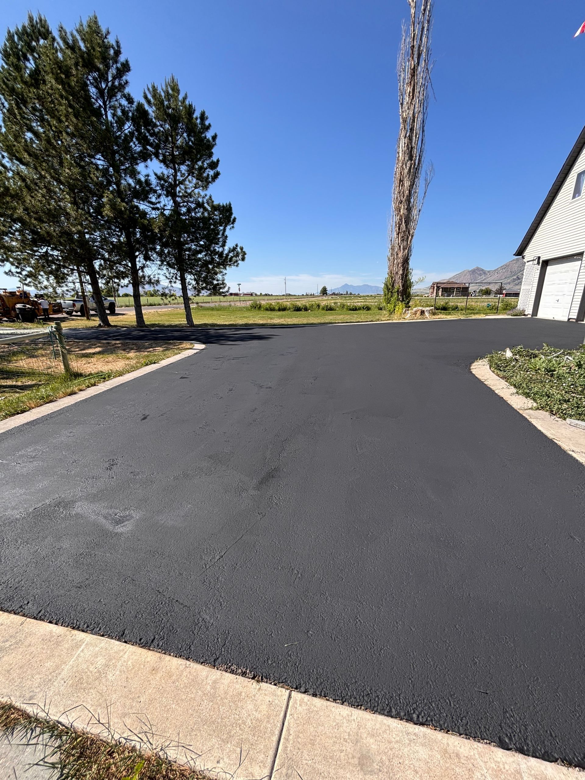 A newly paved black asphalt driveway curves towards a house under a clear blue sky, bordered by concrete and green trees.