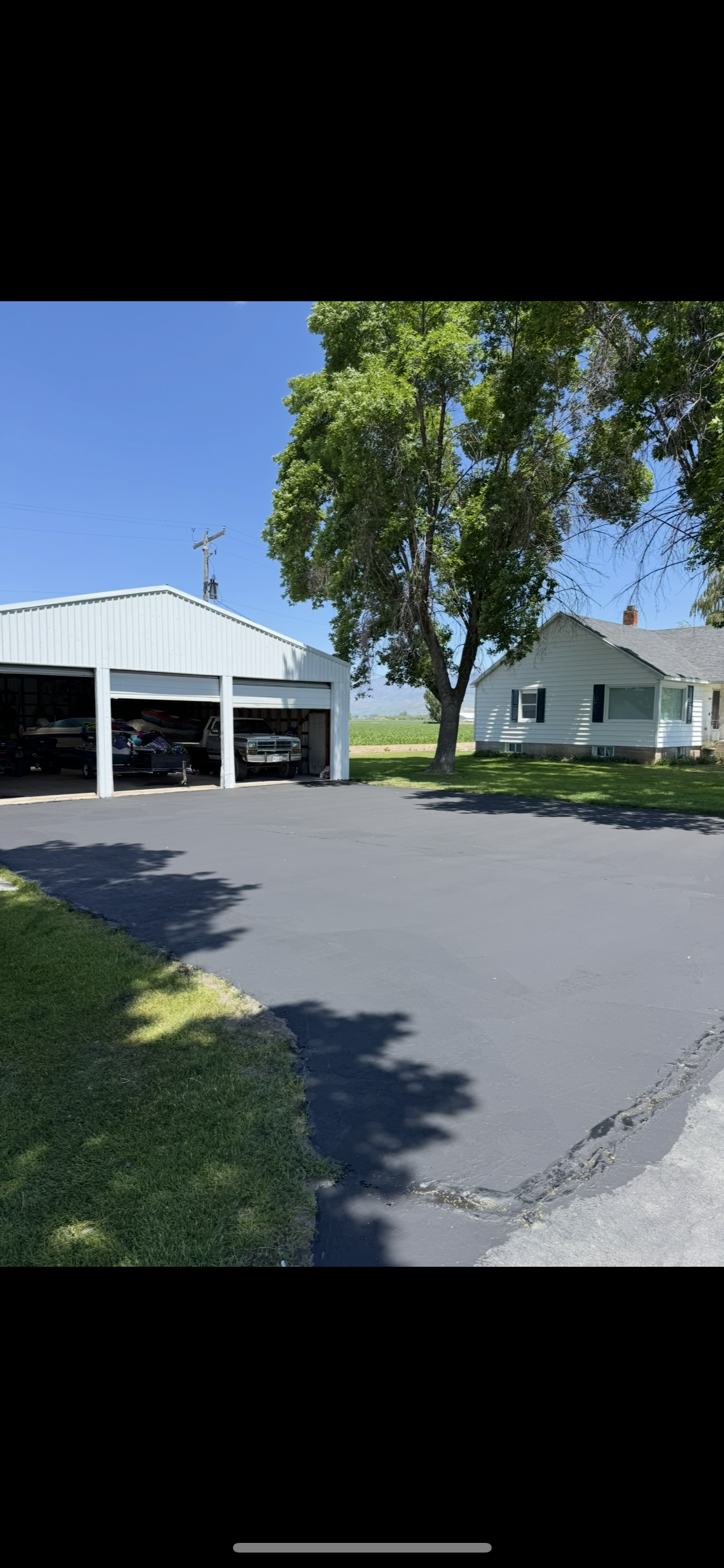A white multi-car garage with an asphalt driveway in front, with a white house and a large tree in the background.