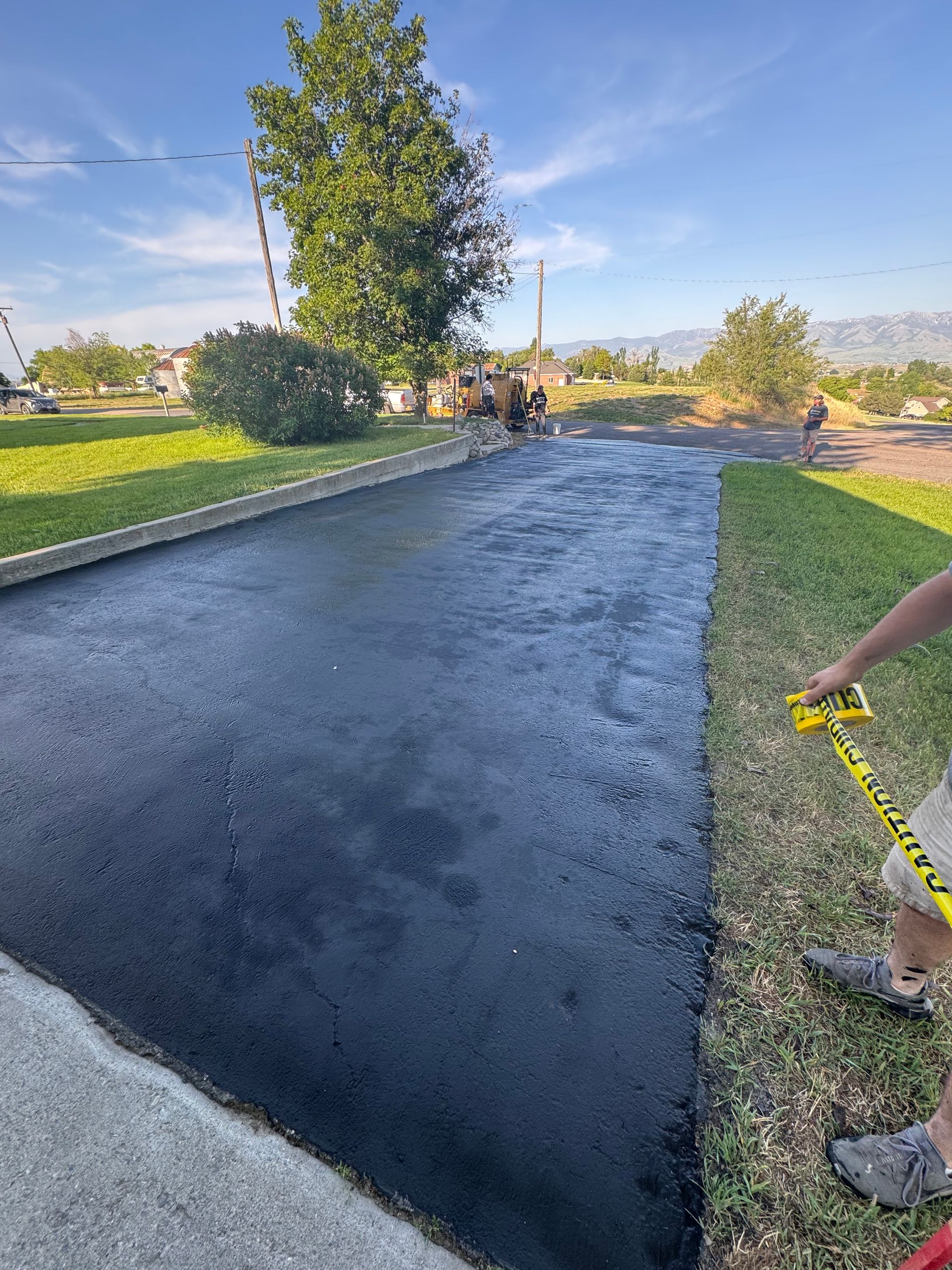 A newly sealed, dark black driveway bordered by green grass under a clear blue sky, with a yellow caution tape dispenser.