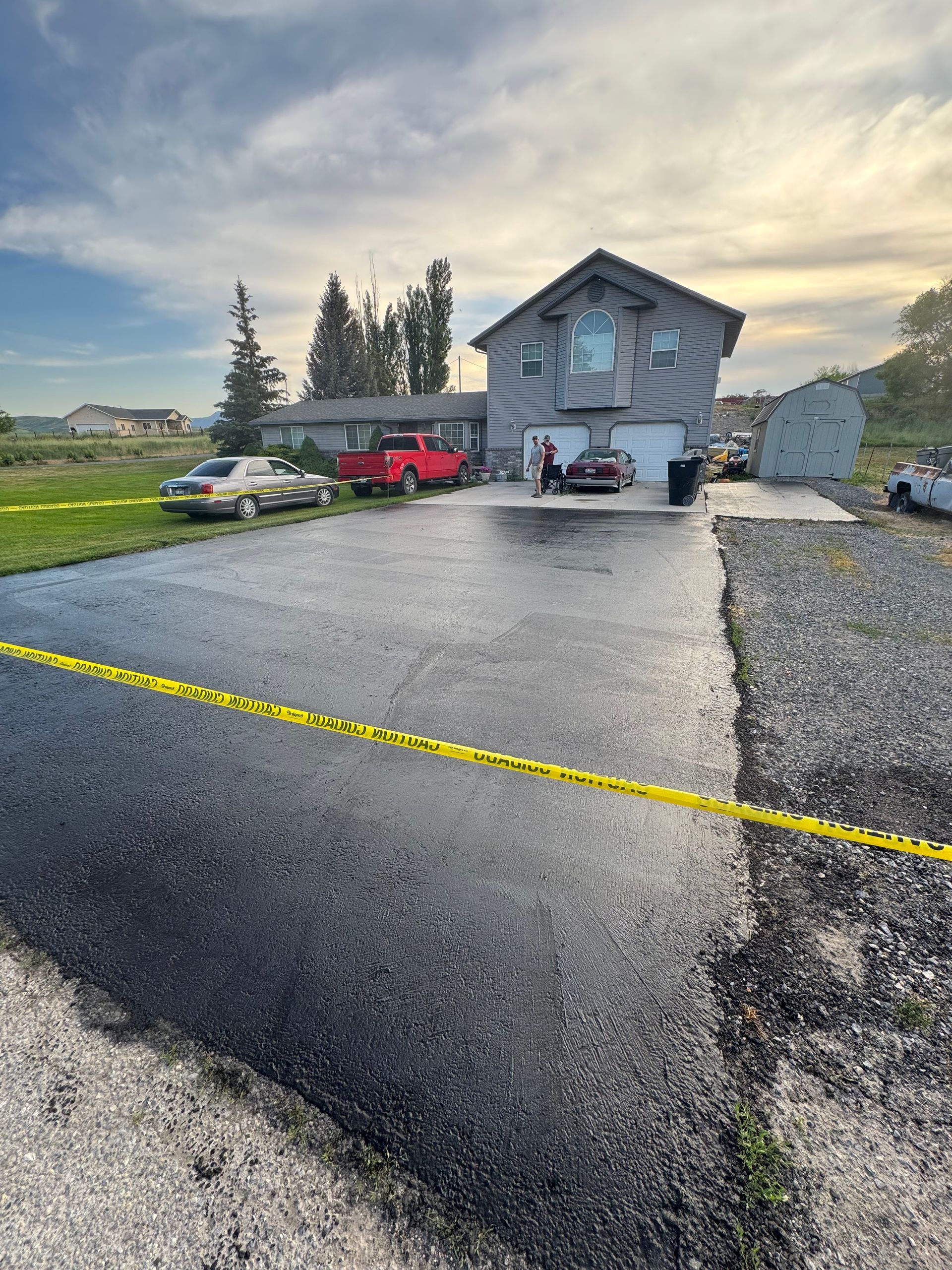 Freshly paved asphalt driveway in front of a gray multi-story suburban home, marked off by yellow caution tape.