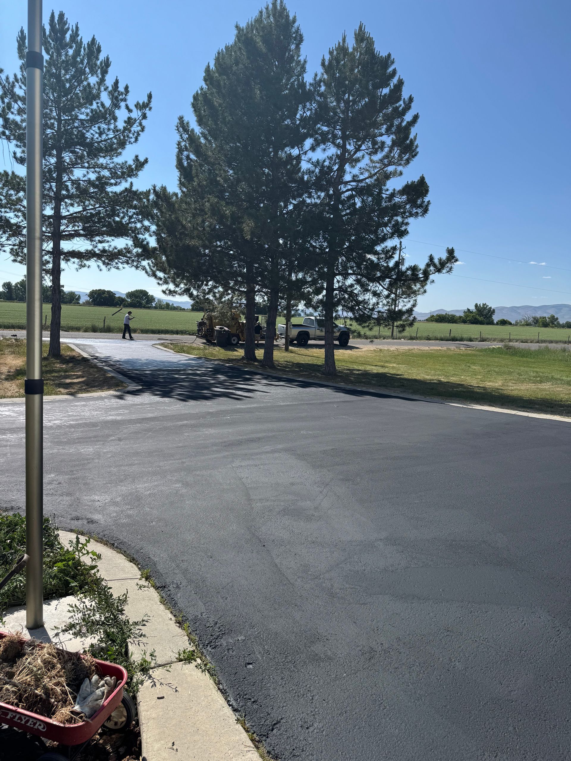 Freshly paved black asphalt driveway leading toward pine trees and a distant vehicle under a bright blue sky.