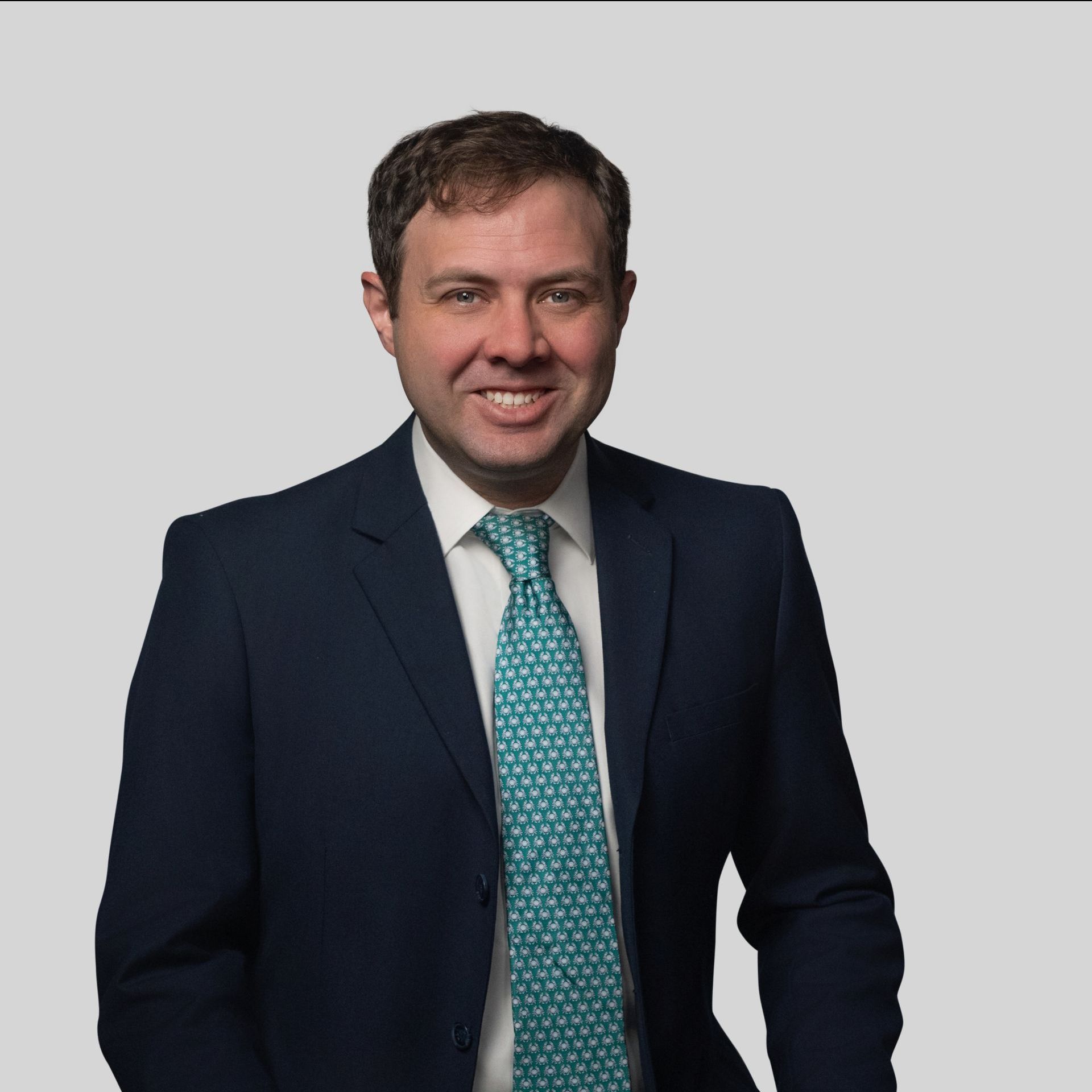 Man in a dark suit and teal tie, smiling against a plain gray background.