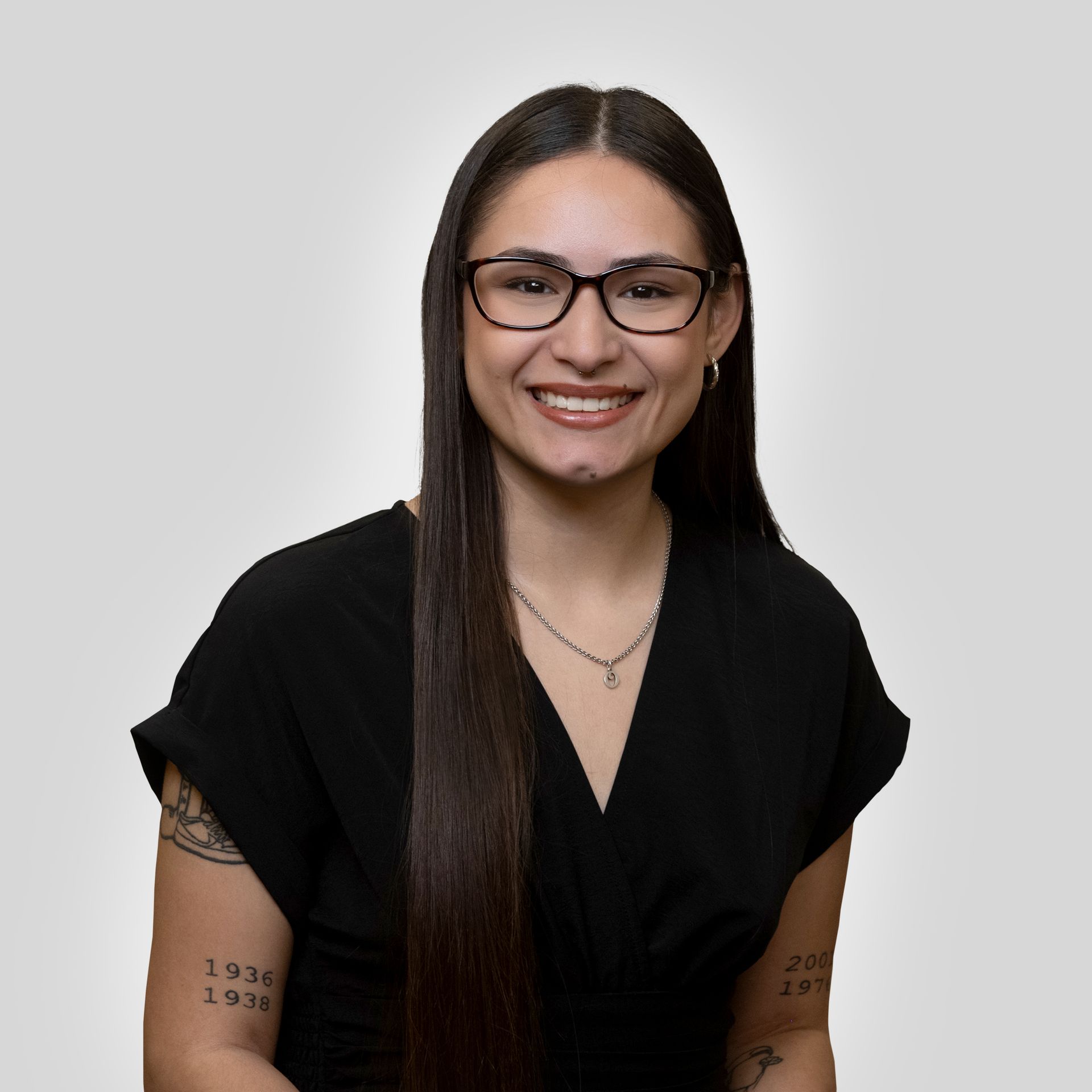 Smiling person with long dark hair and glasses, wearing a black top against a light gray background