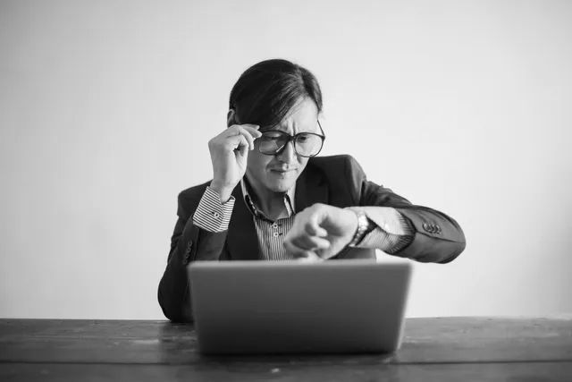 Person looking at a wristwatch while using a laptop at a desk in black and white