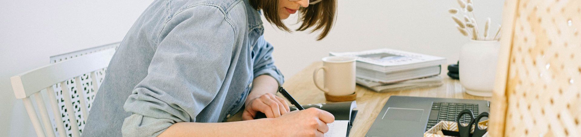 Woman writing at a desk with a laptop, notebook, and stacked books in a bright home office