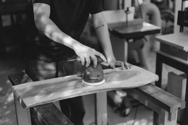 Person sanding a wooden board with an electric sander in a workshop.