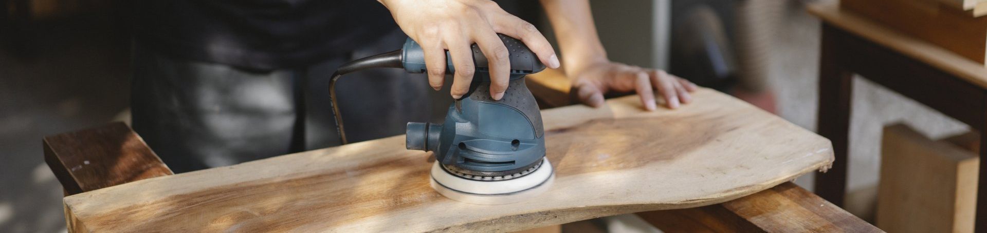 Hands sanding a wooden board with an orbital sander in a workshop