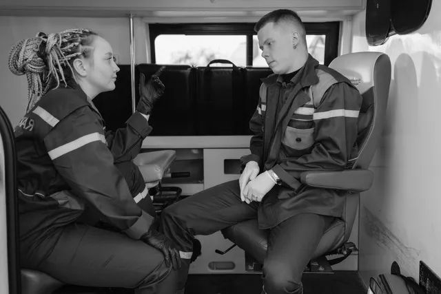 Two rescue workers in dark uniforms sit inside an ambulance, talking beside a stretcher.