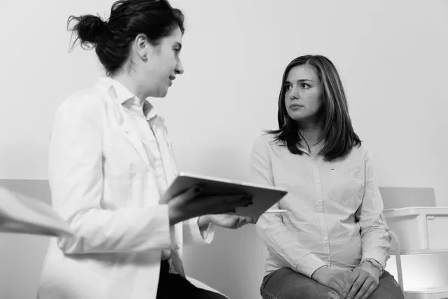 Doctor speaking with a seated patient in a clinic waiting area, holding a clipboard.