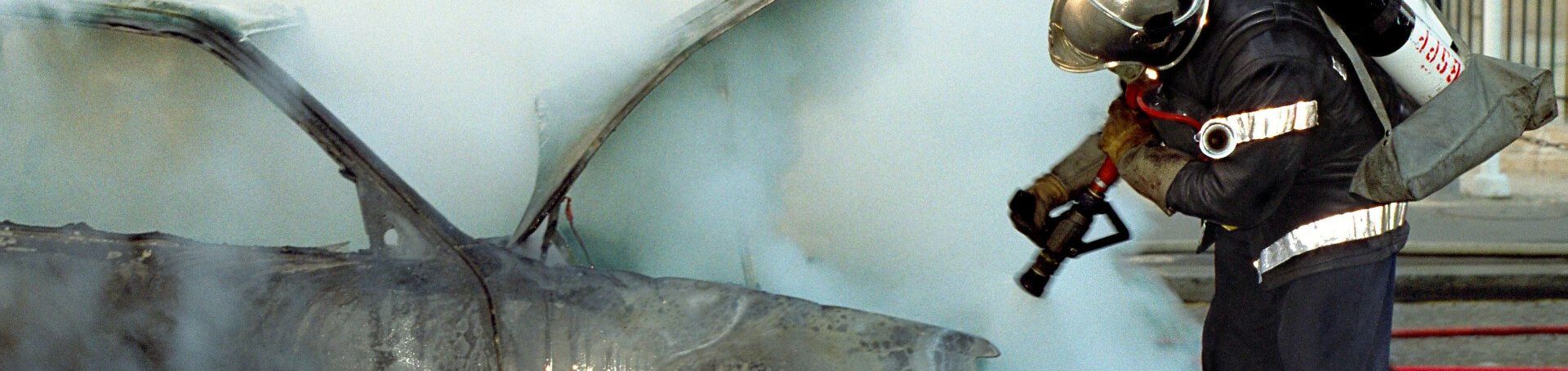 Firefighter in dark gear sprays water on smoking debris during a blaze