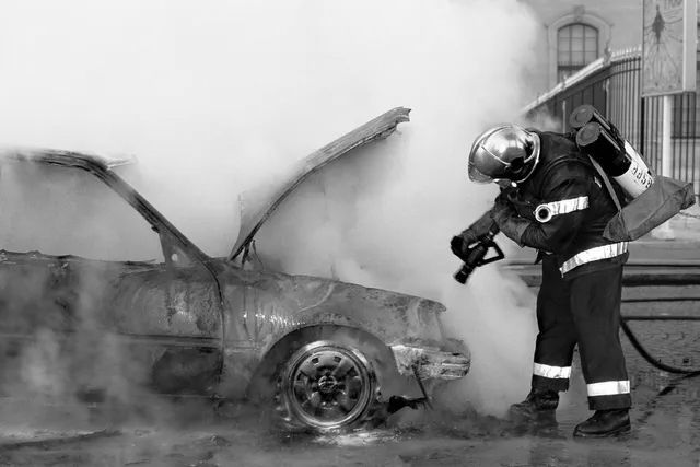 Firefighter spraying water on a smoking car with its hood open beside a building