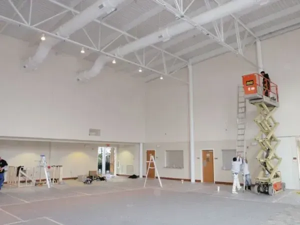 Large empty indoor hall with high white ceiling, ladders, and workers near the right wall