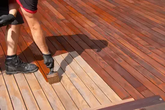 Person staining a wooden deck, brushing a lighter plank section among darker boards.