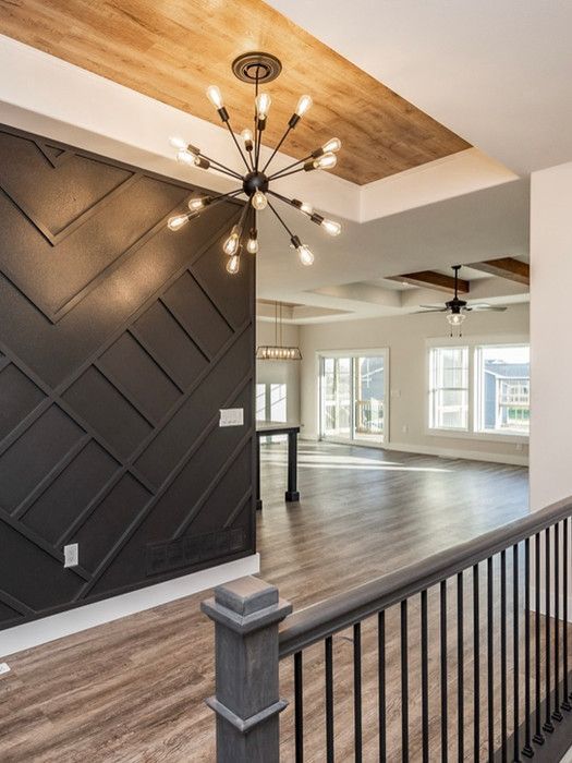 Modern interior with black accent wall, wood ceiling, chandelier, and railing overlooking a bright room.