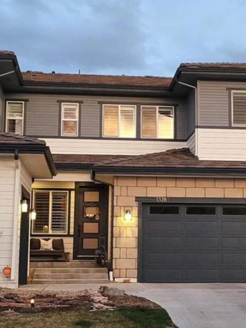 Two-story suburban house with a dark garage door, lit porch, and warm upstairs windows at dusk