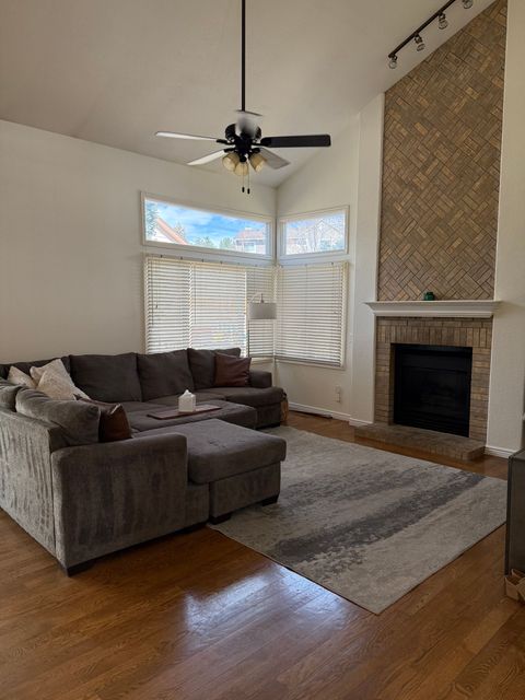 Living room with gray sectional, ceiling fan, fireplace, large windows, and hardwood floors