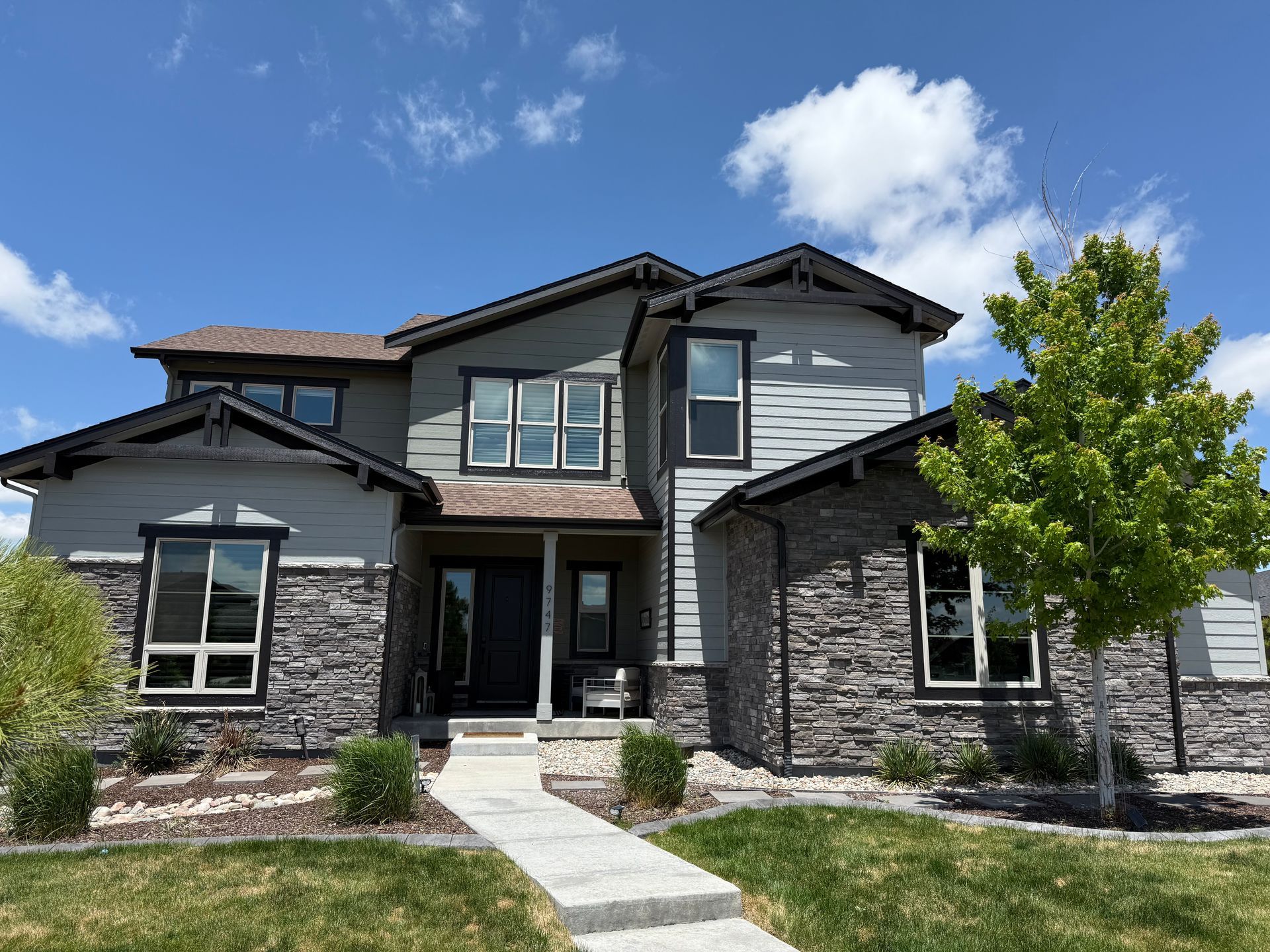 Modern two-story house with stone and gray siding, front porch, and landscaped yard under a blue sky